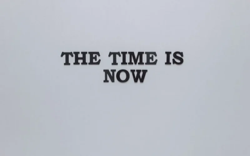 Minimalist poster with the motivational quote 'The Time is Now' on a gray background.