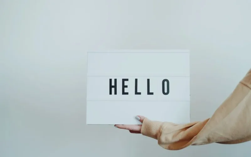 A hand holds a white sign displaying the word 'Hello' against a plain background.