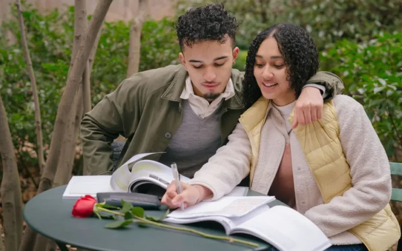 Hispanic boyfriend embracing girlfriend writing in planner at table with fresh red rose in park