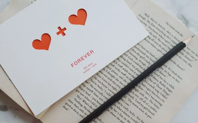 Romantic card with cutout hearts on an open book with a pencil on marble backdrop.