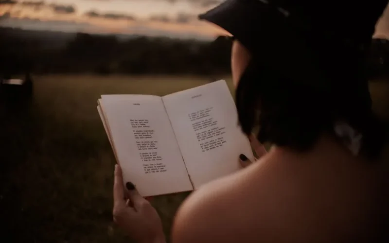 Back view of anonymous female in hat reading book with poetry in grassy field on blurred background on summer evening
