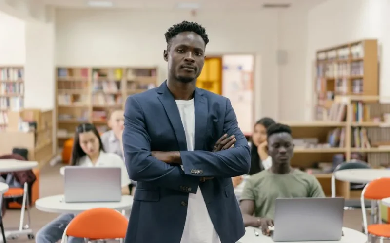 Teacher standing confidently in a library classroom with diverse students using laptops.