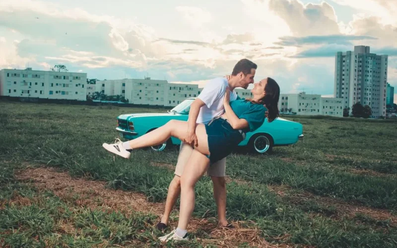 A couple romantically embracing in an open field with a vintage car and buildings in the background.