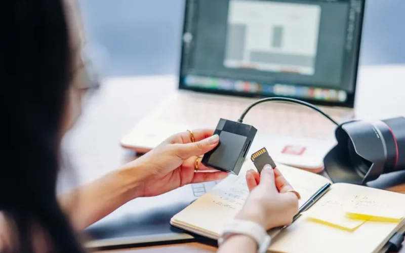 A person prepares to edit photos on a laptop by inserting an SD card, with camera gear on the desk.