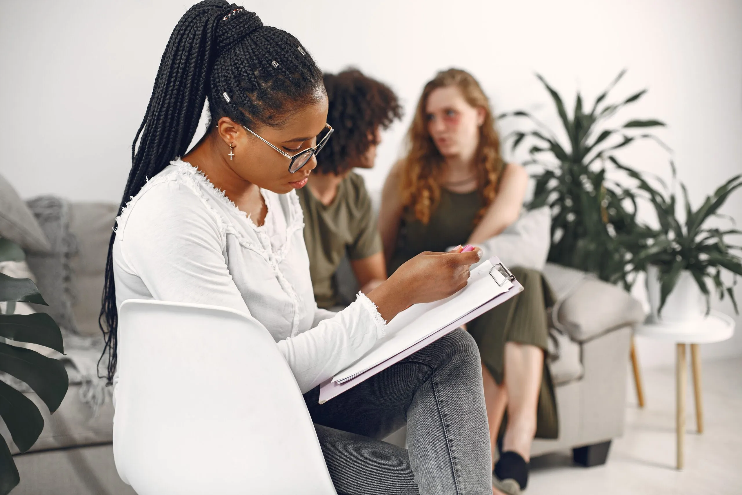 A therapist attentively takes notes while two clients engage in a counseling session indoors.