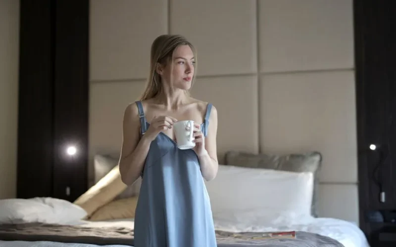 Woman enjoying morning coffee in blue sleepwear, standing in a cozy bedroom setting.