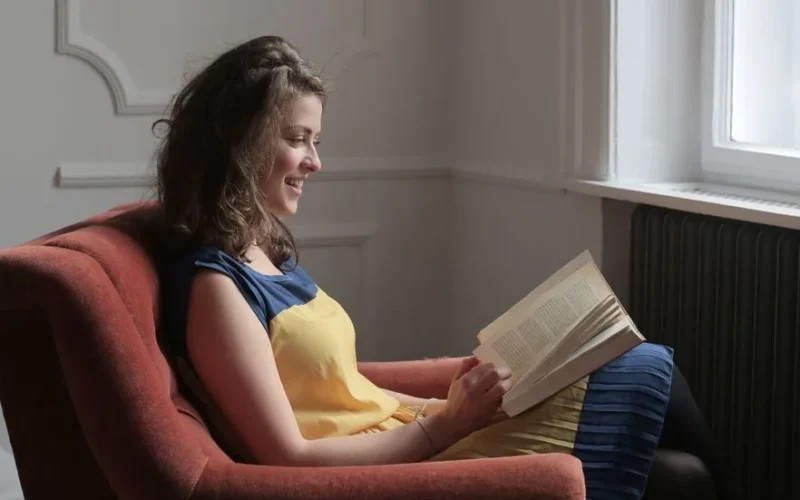 Young woman smiling while reading a book in a cozy chair by a window.