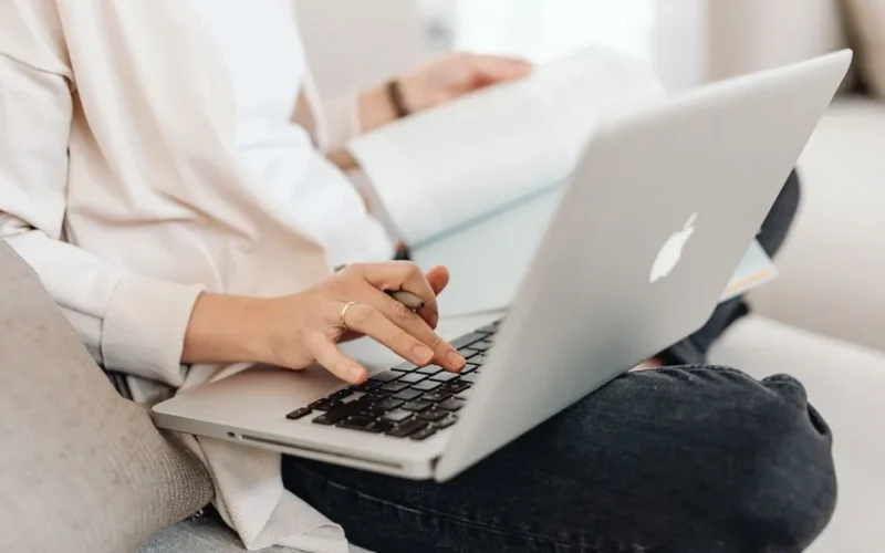 Close-up of a person multitasking, typing on a laptop while holding a book on their lap.