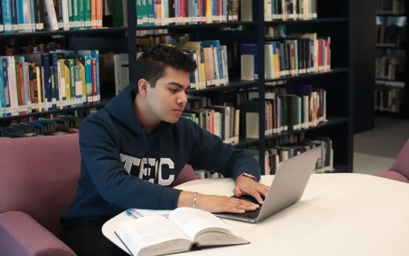 Student in library using laptop and textbooks for study and research.