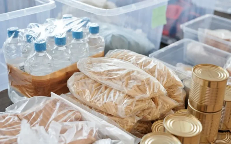 Plastic-wrapped food items and bottled water organized in storage containers and tin cans.