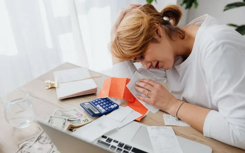 A woman overwhelmed with finances, surrounded by bills, calculator, and cash at home.