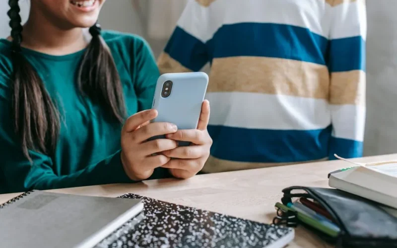 Crop anonymous cheerful little schoolgirl in casual clothes sitting at table in classroom and sharing video on smartphone with unrecognizable friend