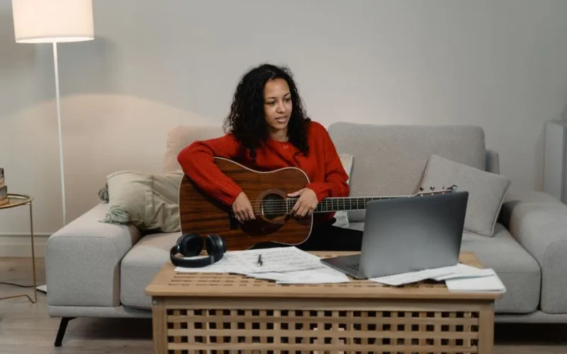 Woman in living room playing guitar while learning online via laptop. Cozy and creative setup.