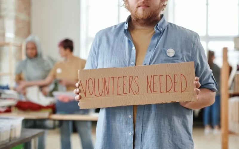 A man holds a 'Volunteers Needed' sign in a charitable organization setting, highlighting the need for help.