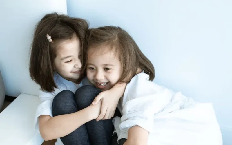 Two young girls in white tops embrace joyfully indoors, capturing sibling love and happiness.