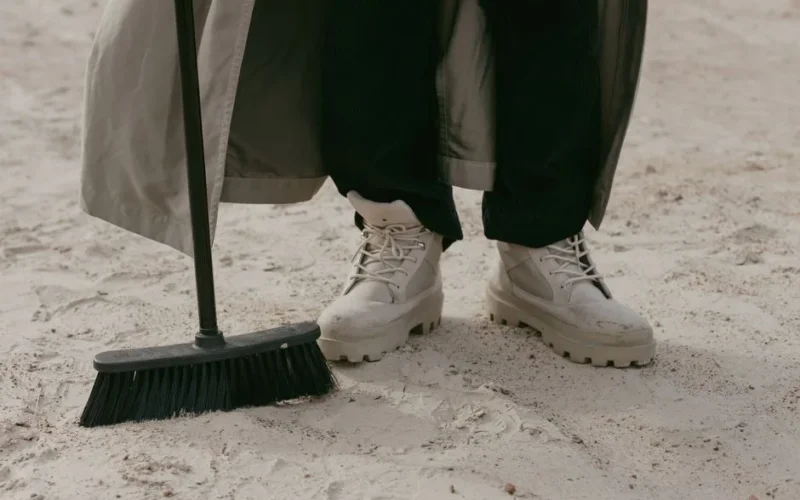 Close-up of a person wearing boots using a broom on sandy terrain, emphasizing footwear style and casual chores.