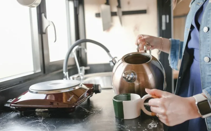 A woman in a denim jacket pours tea from a kettle into a cup on a kitchen counter.