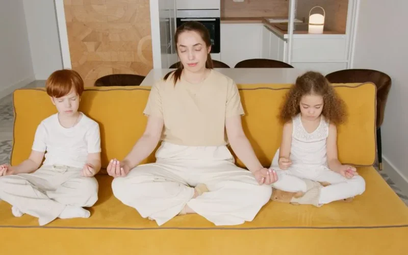 A mother and two children practicing mindfulness meditation indoors on a cozy sofa.