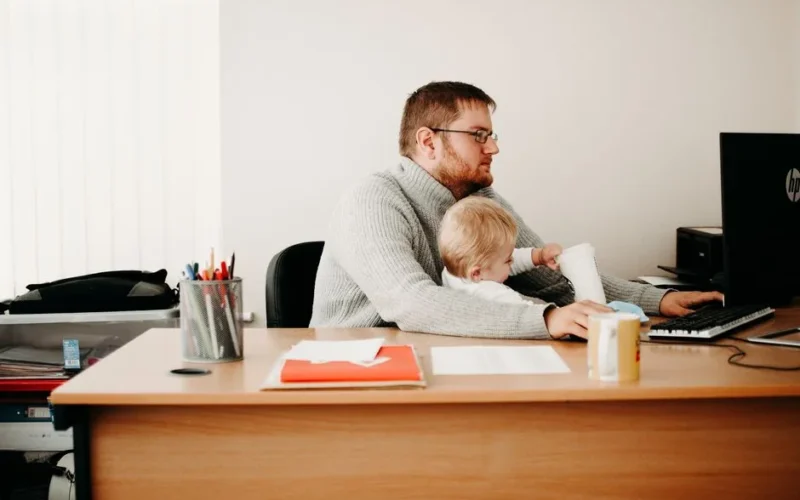 A father multitasks at his desk while holding his child, illustrating remote work and parenting.