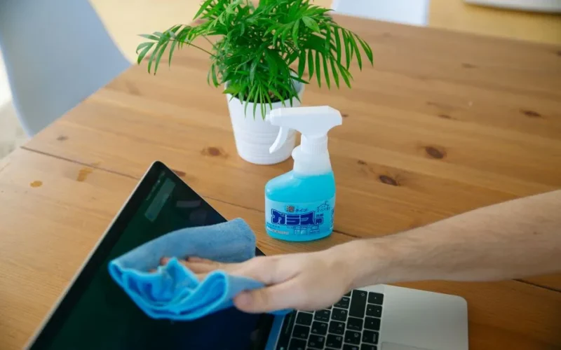 Close-up of hand cleaning a laptop screen with a blue cloth and spray bottle on wooden desk.