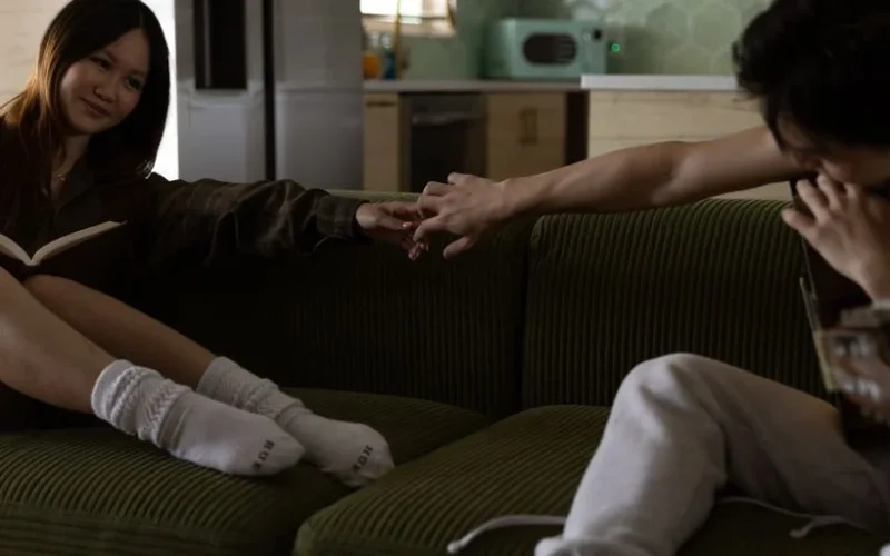 A couple sitting on a couch, holding hands while enjoying a book and playing guitar.
