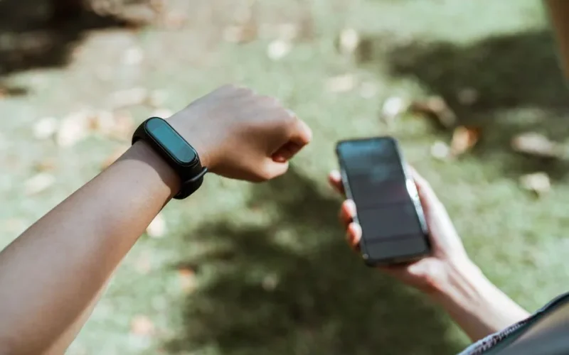 Top view of crop anonymous person demonstrating modern plastic bracelet with small display while surfing internet on cellphone sitting in tranquil park covered with faded leaves in daylight