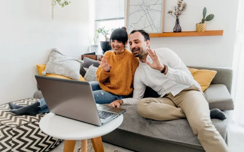 Happy couple sitting on couch, using laptop for a video call in a stylish living room.