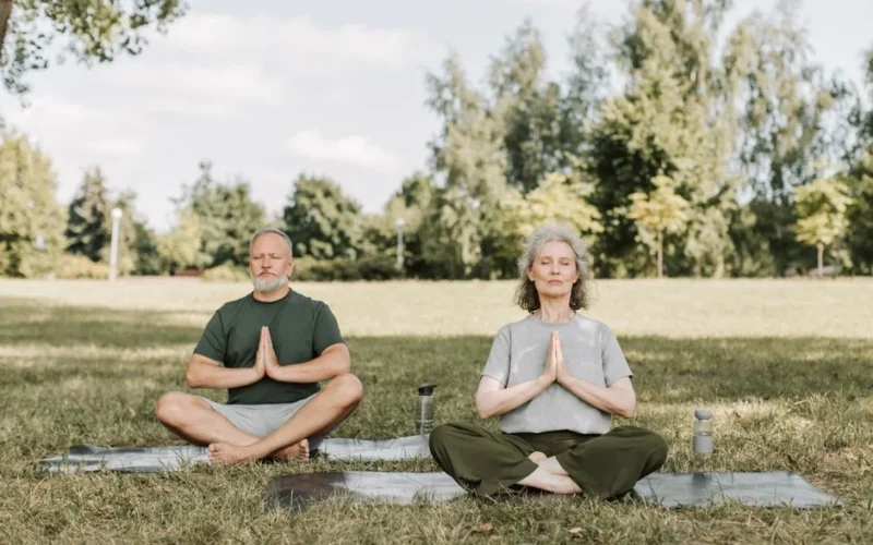 Senior couple meditating on yoga mats in a serene park setting, enjoying peaceful yoga practice.