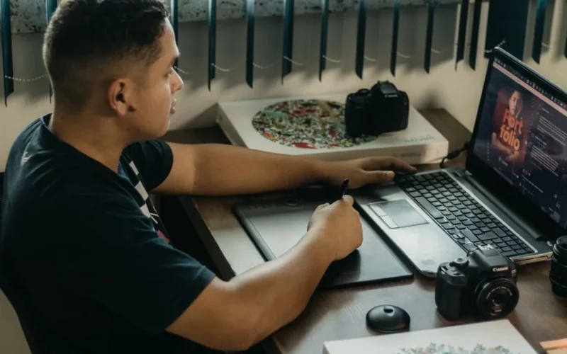 Man editing photos on a laptop using a graphics tablet, set in an indoor workspace with camera equipment.
