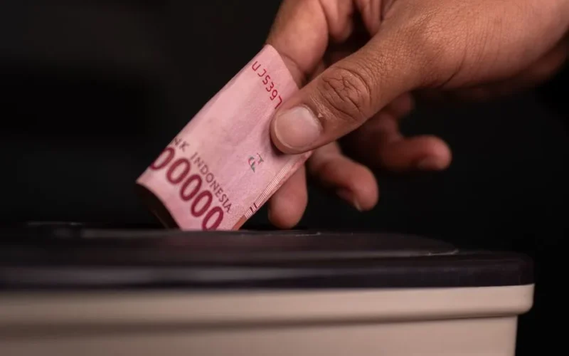 Close-up of a hand donating an Indonesian Rupiah note into a charity box, symbolizing generosity.