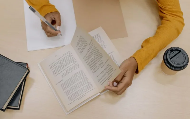 A person writing and reading a book at a desk with a coffee cup nearby.