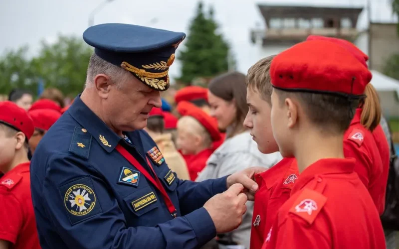 A Russian officer in uniform pinning a badge on a young recruit during an outdoor military ceremony.