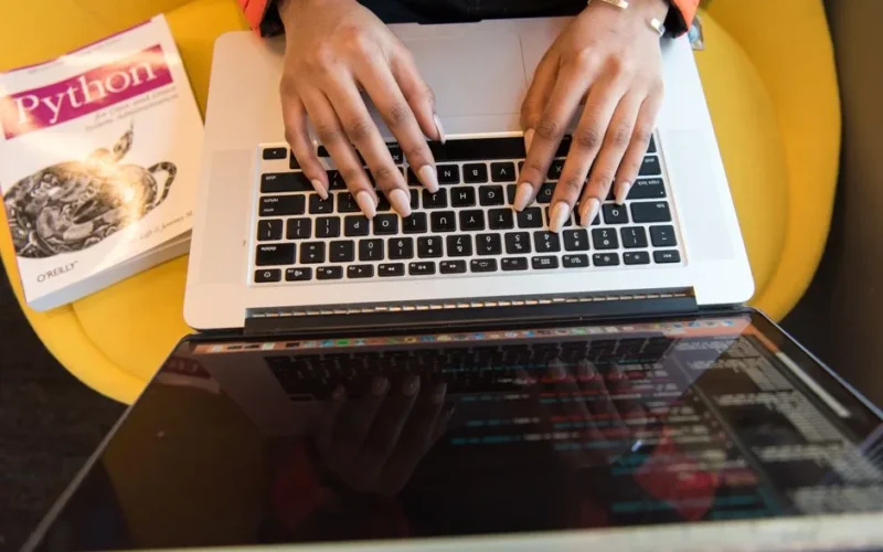 Close-up of hands typing on a laptop keyboard, Python book in sight, coding in progress.