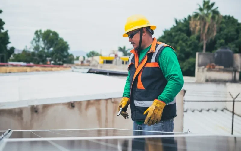 A solar technician in protective gear inspecting rooftop solar panels outdoors.
