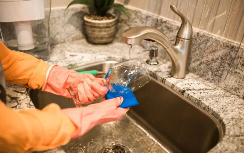 Close-up of hands wearing gloves washing a glass in a kitchen sink under a running faucet.