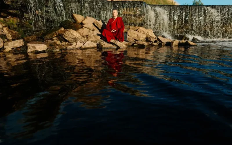 Monk in red robe meditating by a tranquil waterfall, promoting peace and serenity.
