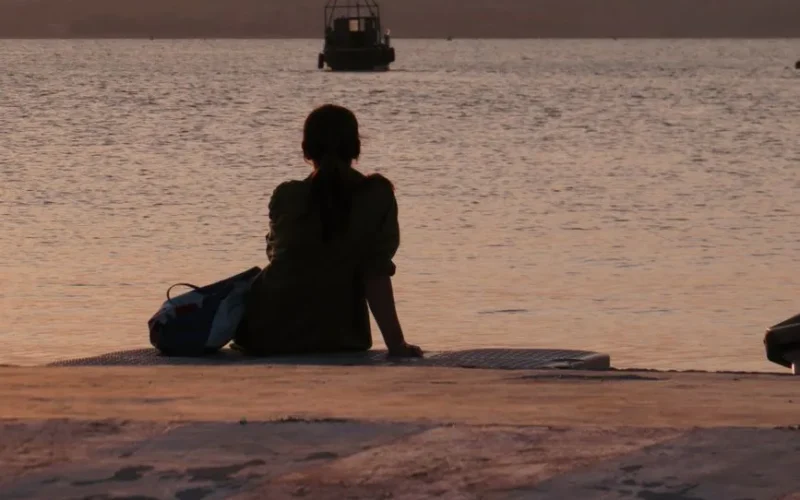 A woman sits by the sea at sunset, gazing at the horizon in peaceful solitude.