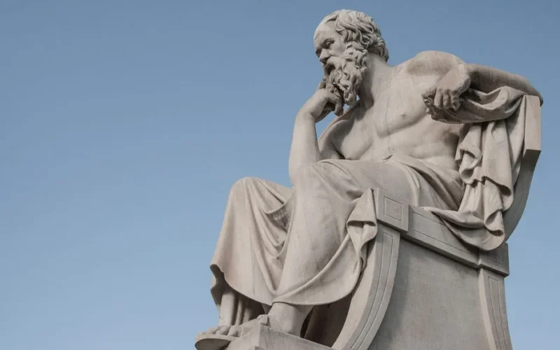 Stone statue of philosopher Socrates sitting in thoughtful pose in Athens, Greece under a clear blue sky.