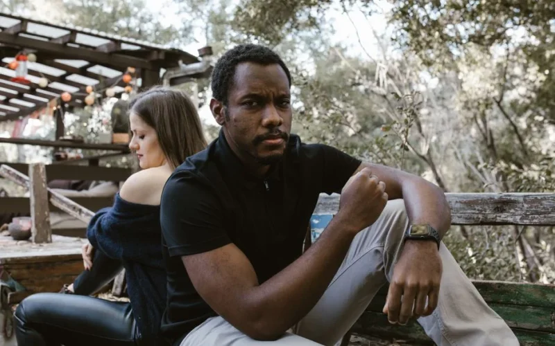 A thoughtful man sits outdoors, with a woman sitting distantly behind him.