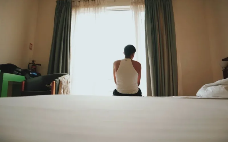 A man sits indoors facing a bright window, lost in thought, casting an introspective mood.