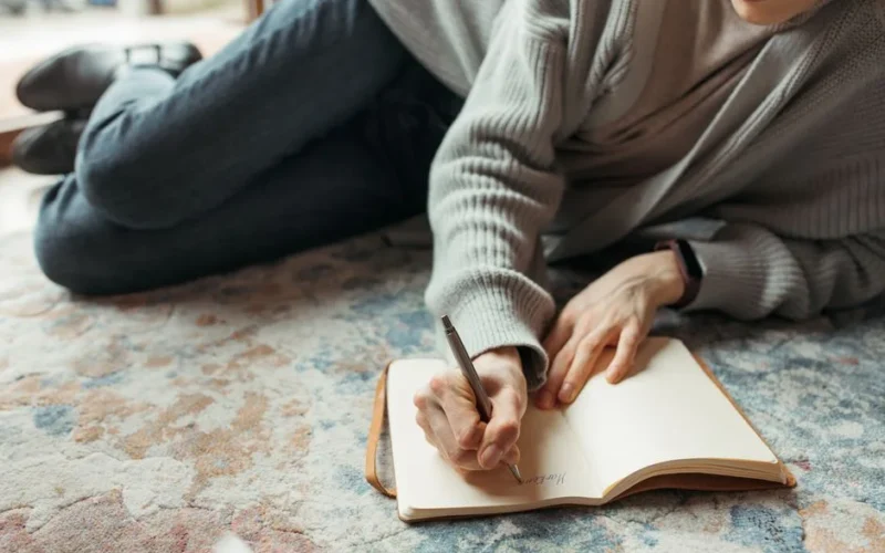 A person lies comfortably on the floor, writing in a journal with a pen, symbolizing creativity and relaxation.