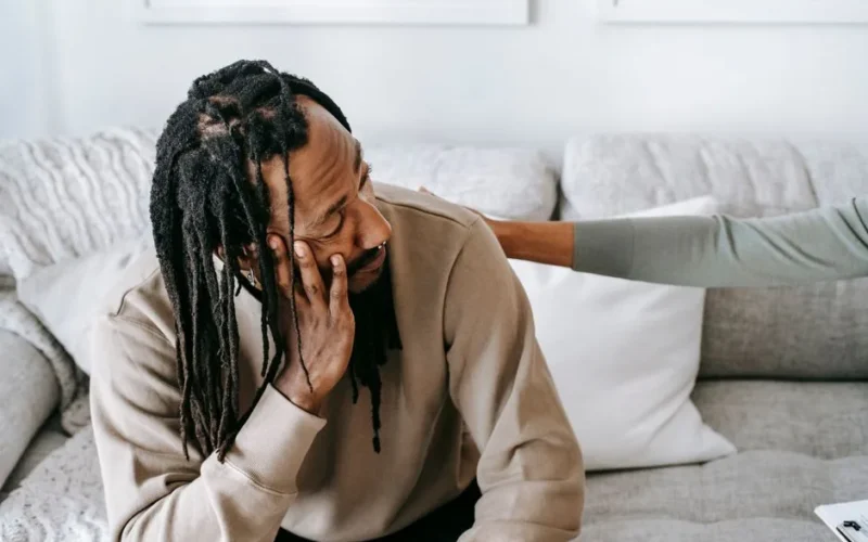 Depressed African American male in uneasy situation discussing problem with female psychologist or friend while sitting on sofa together