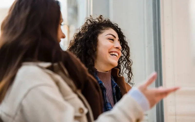 Side view of happy young multiethnic female best friends with long dark hairs in casual clothes laughing while looking at showcase of fashion store on city street