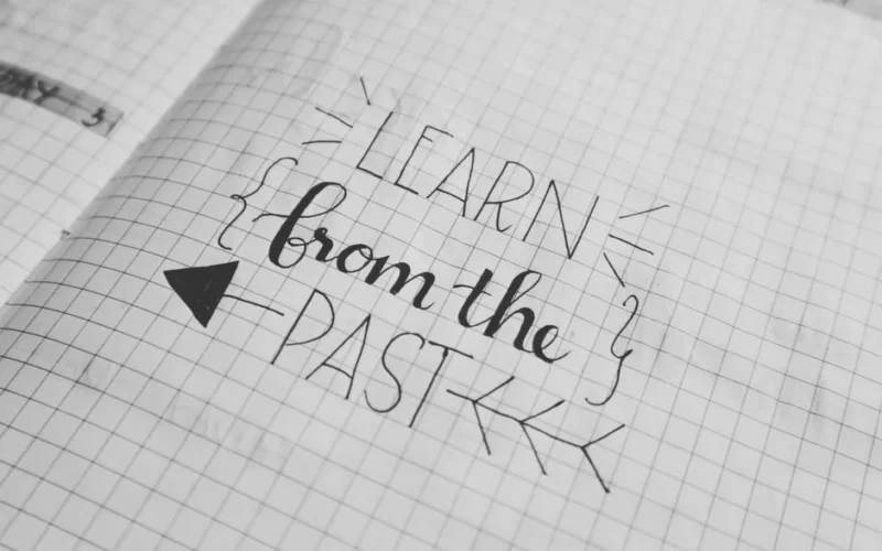 Black and white close-up of a handwritten motivational quote 'Learn from the Past' on graph notebook paper.