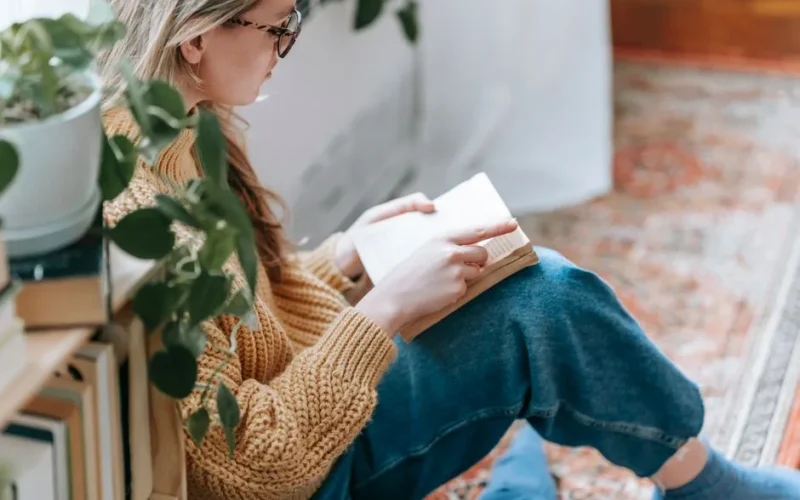 Side view of crop female student in casual clothes sitting on carpet near bookshelf with plant and reading book at home