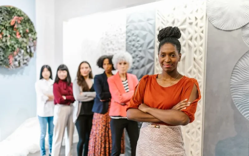 A confident female leader stands with diverse female colleagues in a modern office setting.