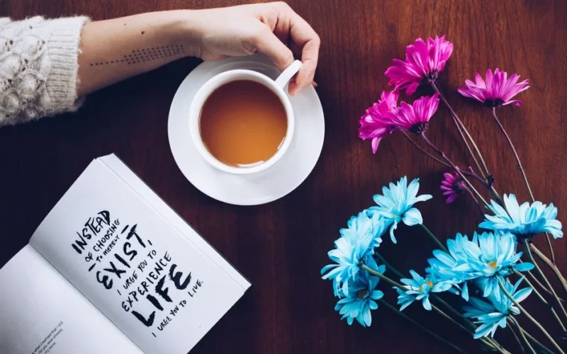 A cozy setup with tea, flowers, and an inspirational quote book on a wooden table.