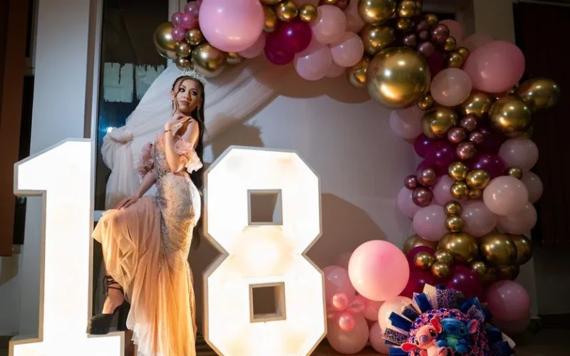 A young woman poses by illuminated 18, surrounded by colorful balloons in a festive indoor setting.