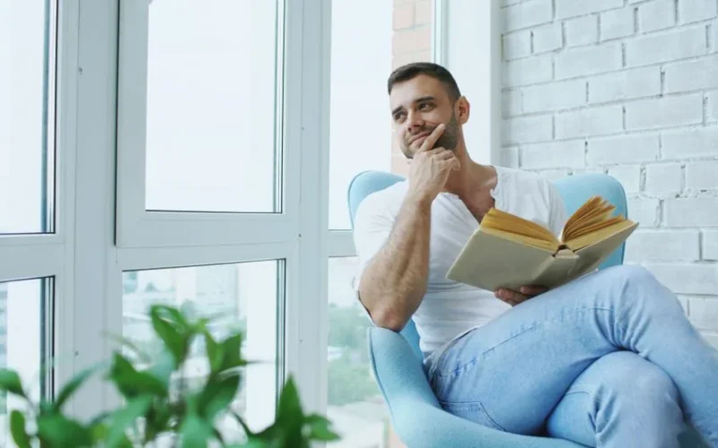 Thoughtful man sitting in chair reading a book by the window