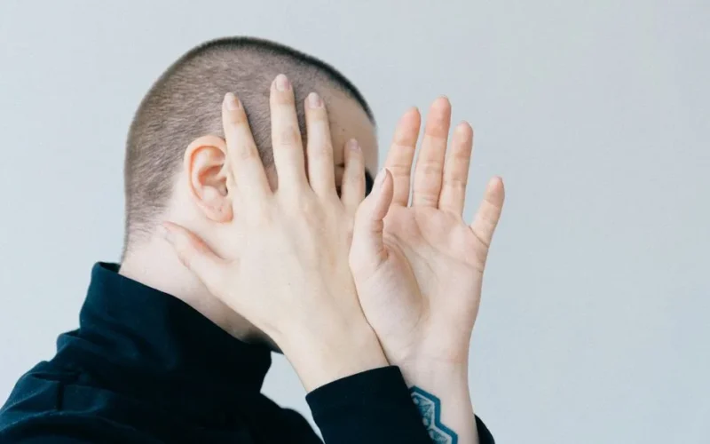 A person hides their face with hands in a minimalist portrait with a light background.